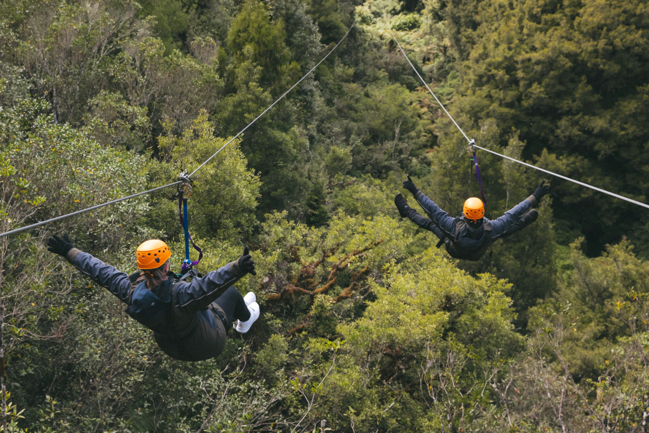 Two people wearing orange helmets are suspended on parallel zip lines over a dense green canopy.